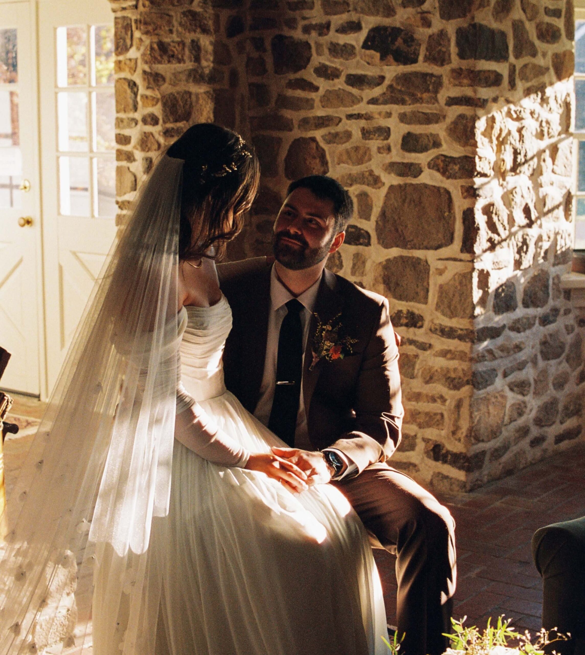 A bride in a white wedding dress and veil sits on the lap of a groom in a brown suit in a sunlit stone-walled room.