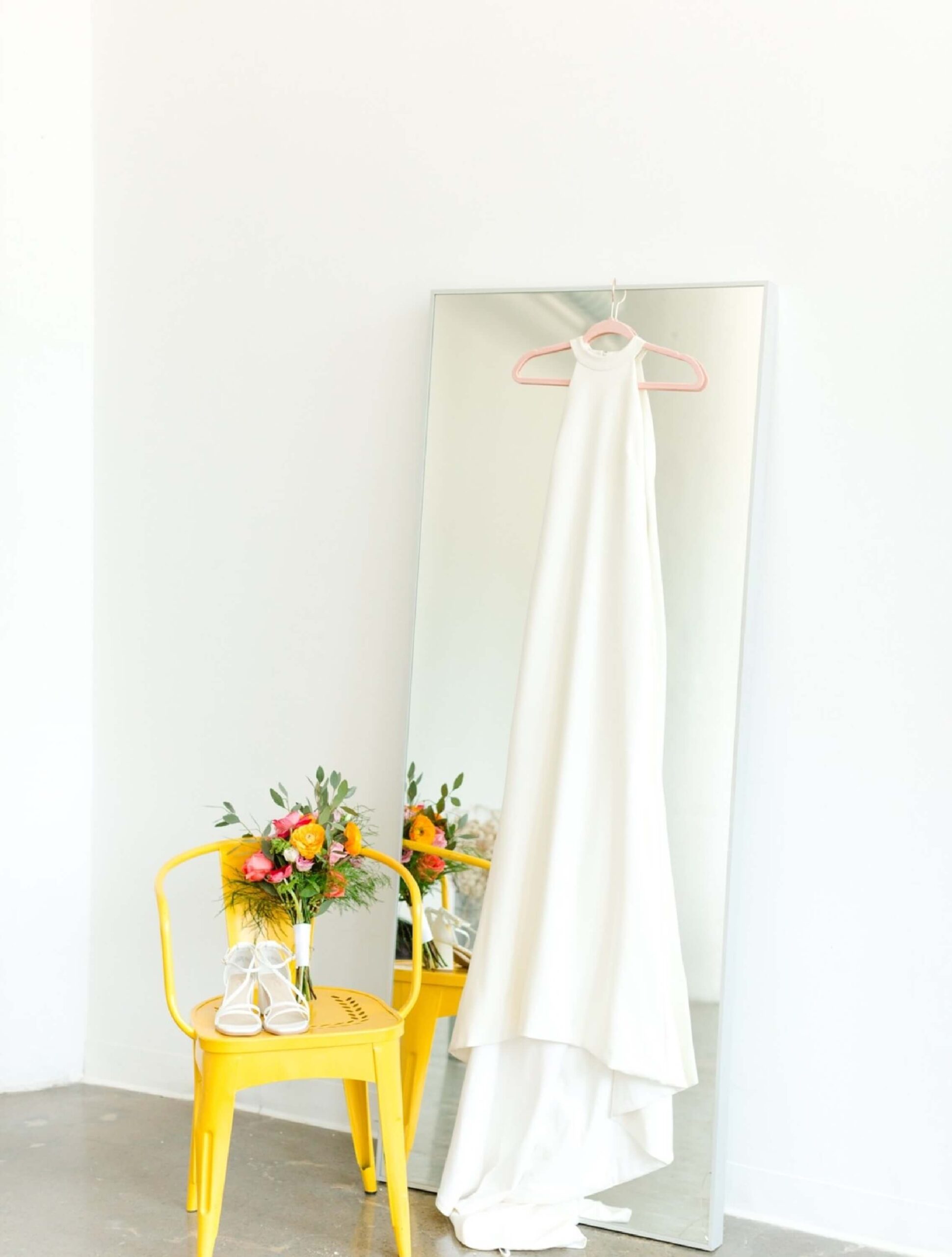 A white sleeveless gown hangs on a mirror next to a yellow chair holding a bright floral bouquet and a pair of white shoes.