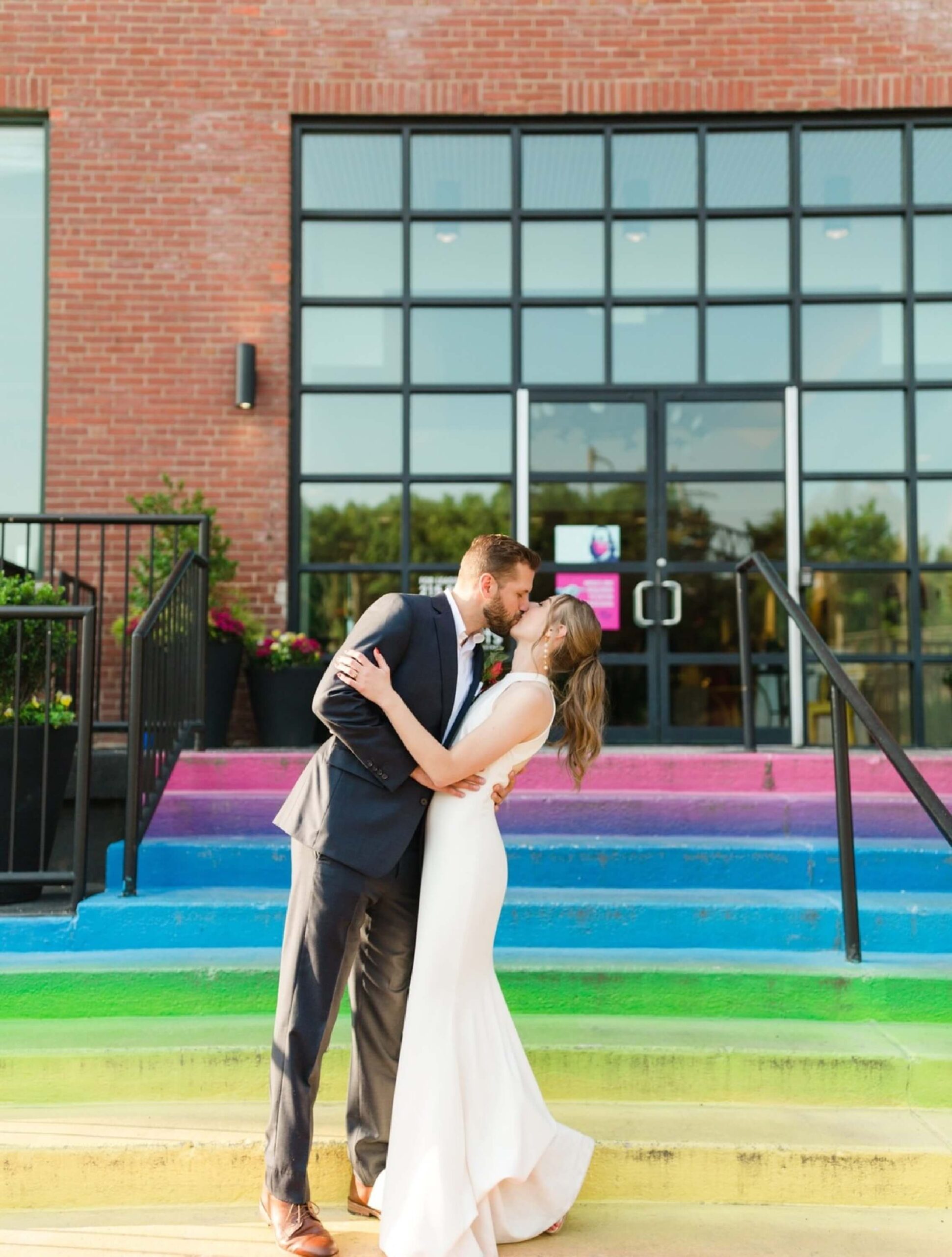 A couple dressed in wedding attire kiss on colorful rainbow steps in front of a modern brick building with large glass doors.