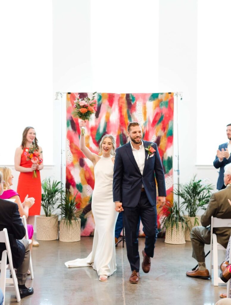 A bride and groom walk down the aisle, smiling and holding hands, as guests applaud in a brightly lit room with a colorful backdrop.