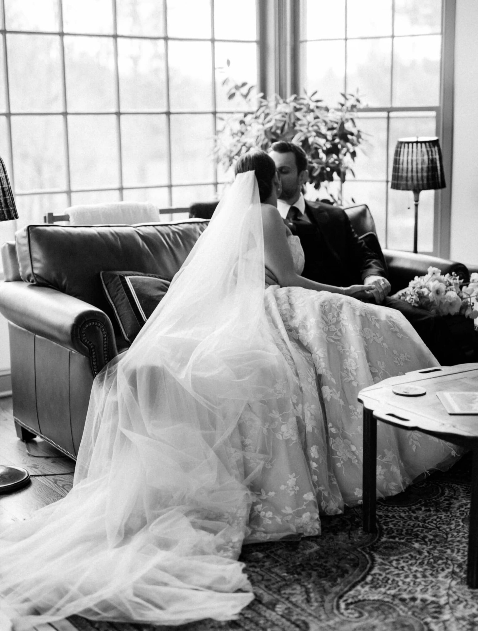 A bride and groom sit on a couch and kiss, with the bride wearing a long veil and floral gown, in a room with large windows and soft lighting.