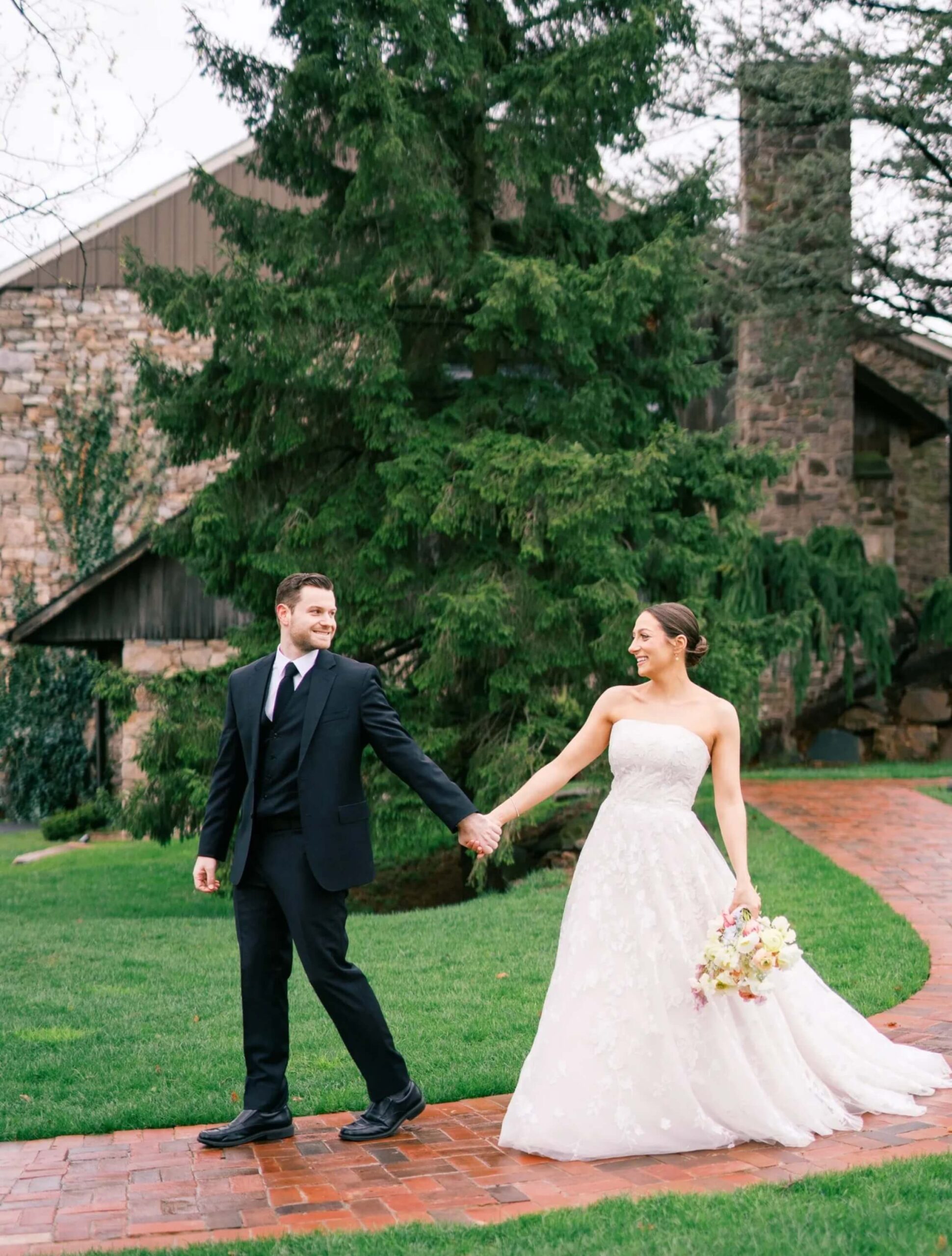 A bride in a white gown holds a bouquet and the hand of a groom in a black suit as they walk on a brick path outside near a large evergreen tree and stone building.