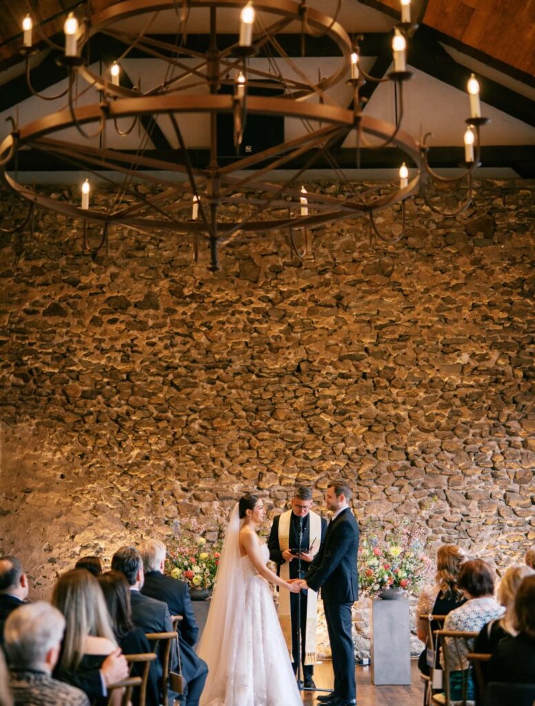 A bride and groom stand before an officiant holding hands during an indoor wedding ceremony, with guests seated and a stone wall in the background.