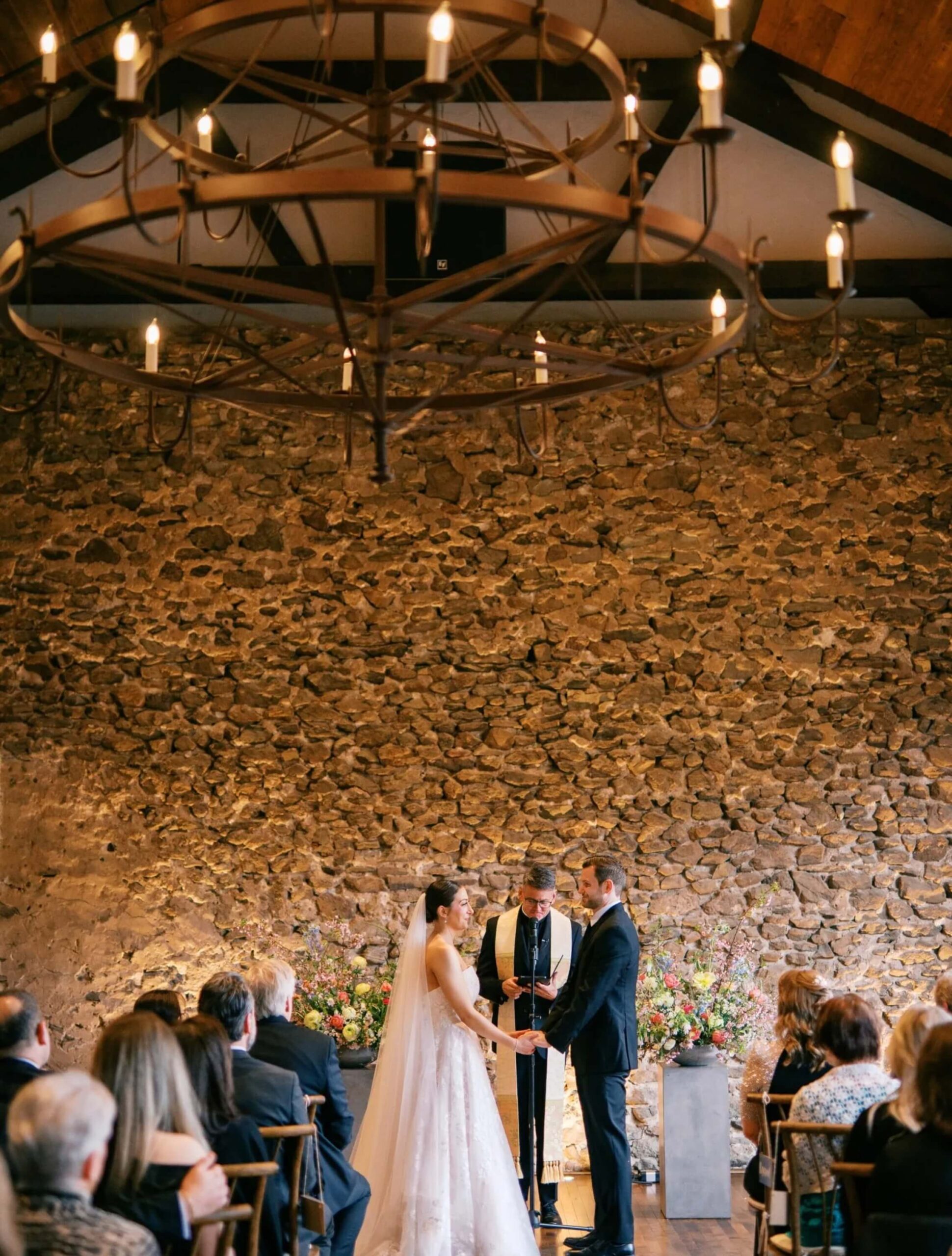 A bride and groom stand before an officiant holding hands during an indoor wedding ceremony, with guests seated and a stone wall in the background.