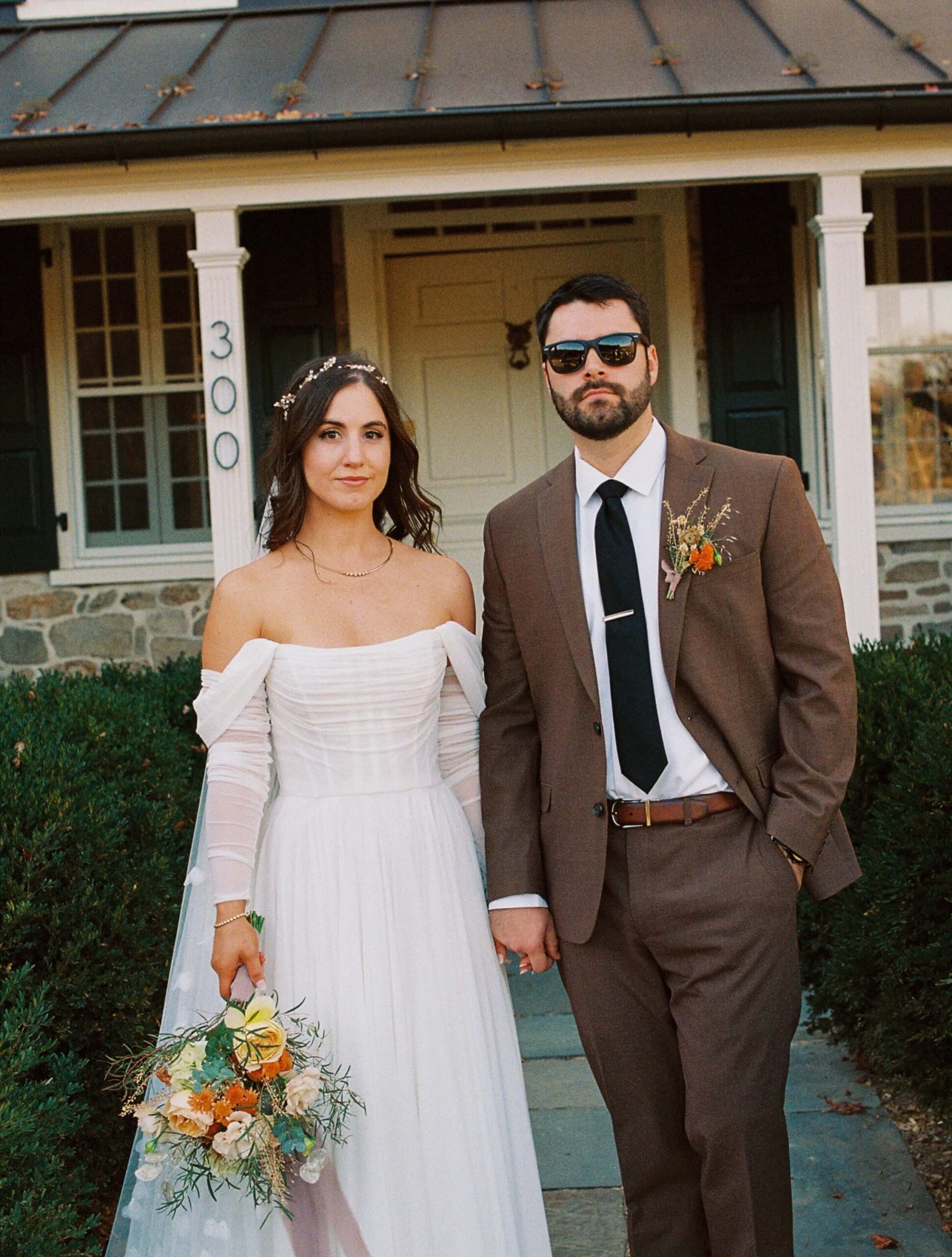 A bride in a white dress and a groom in a brown suit and sunglasses stand holding hands outside a stone house.