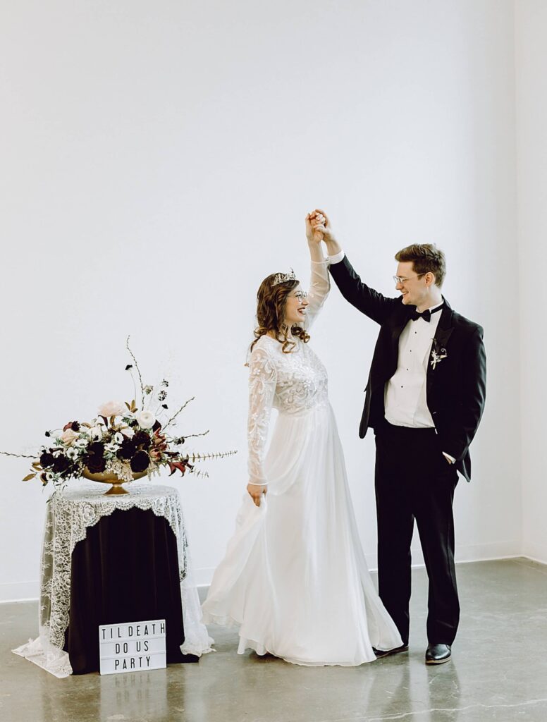 A bride and groom dance together in formal attire next to a flower arrangement and a sign that reads 