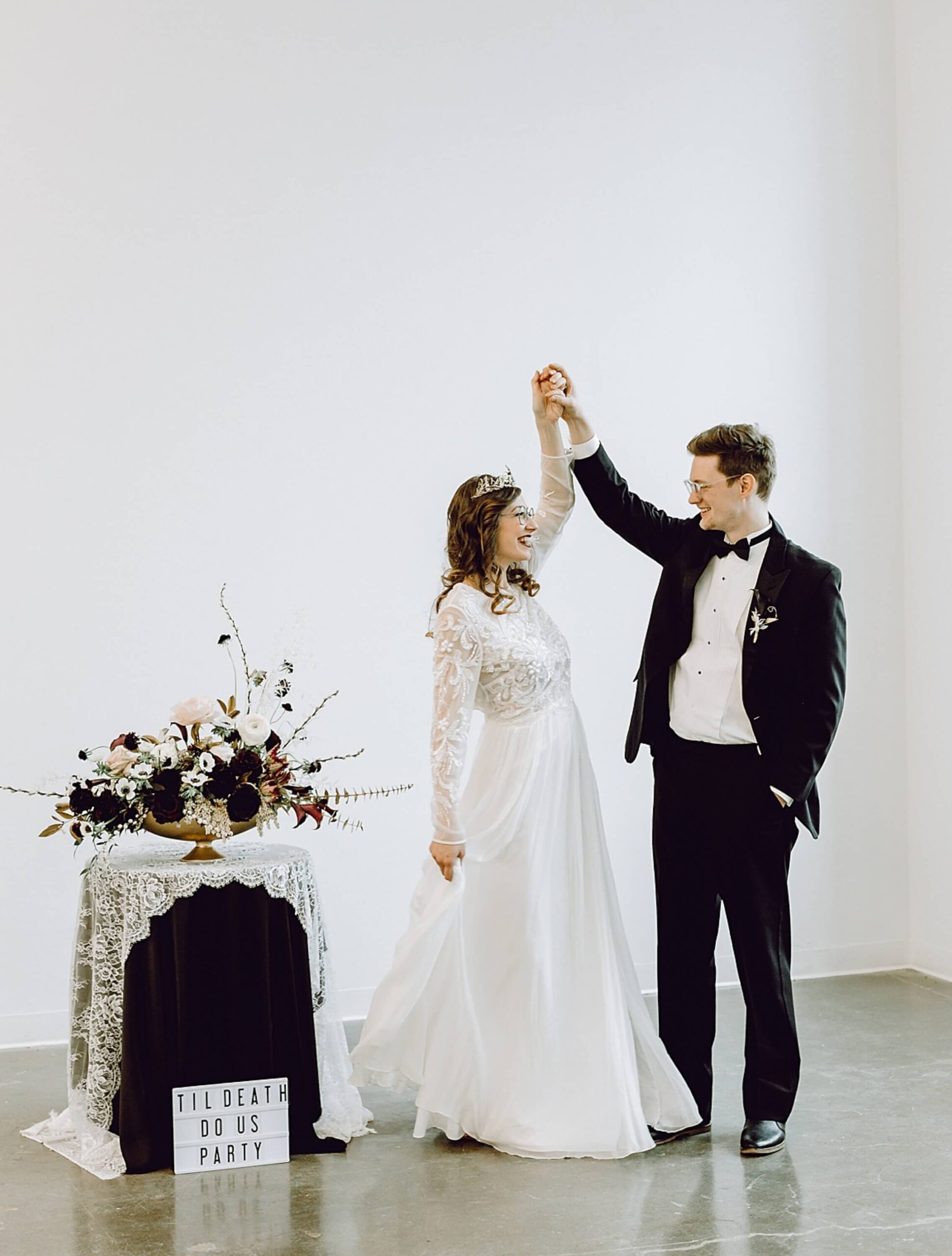 A bride and groom dance together in formal attire next to a flower arrangement and a sign that reads 