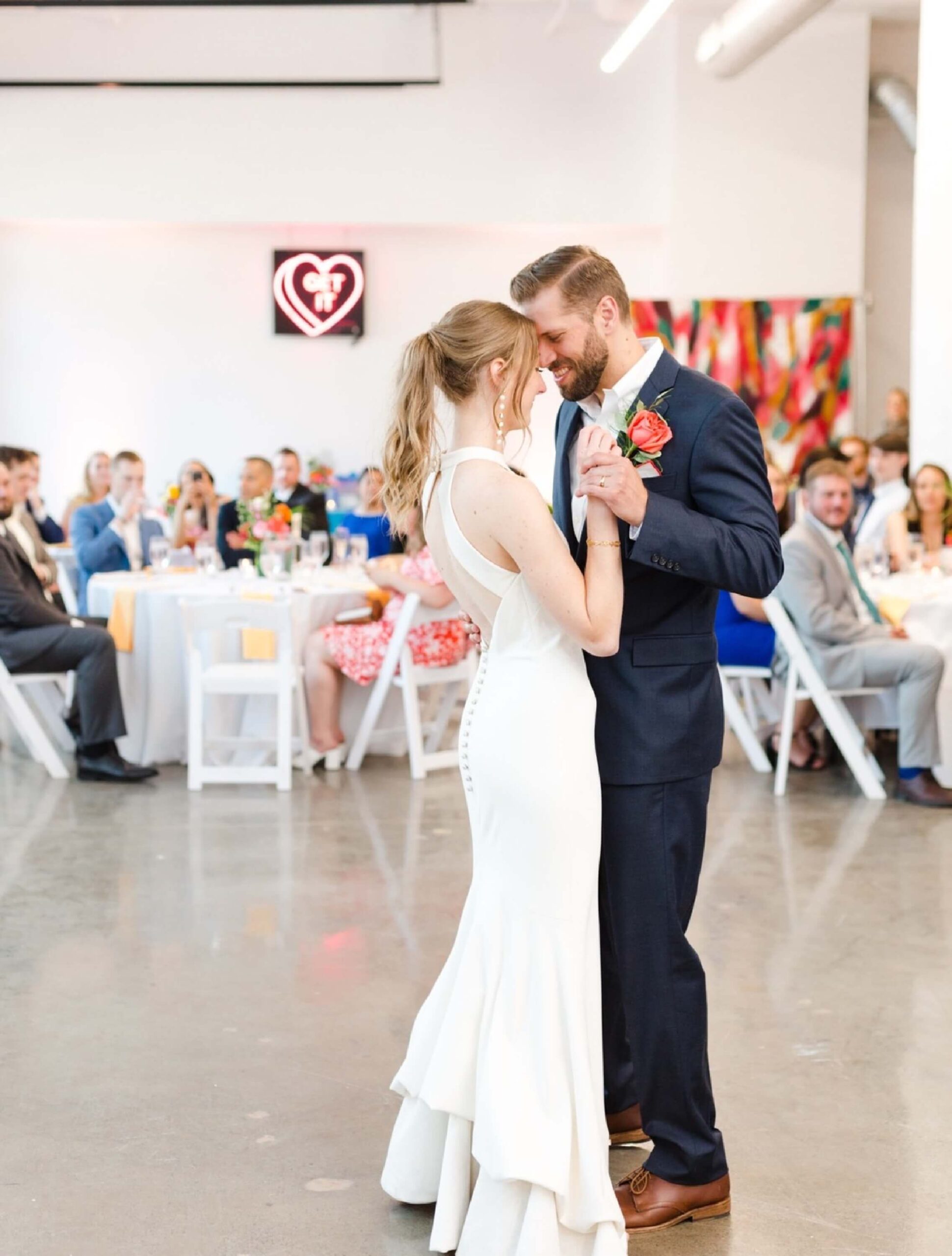 A bride and groom share their first dance in a brightly lit reception hall, with guests seated at decorated tables in the background.