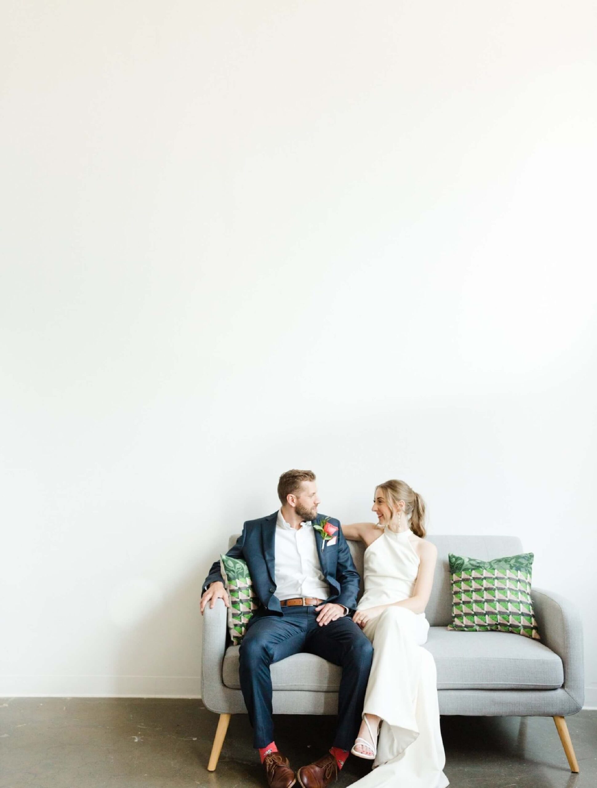 A bride and groom sit together on a gray couch with green patterned pillows, facing each other in a bright, minimalist room with a white wall.