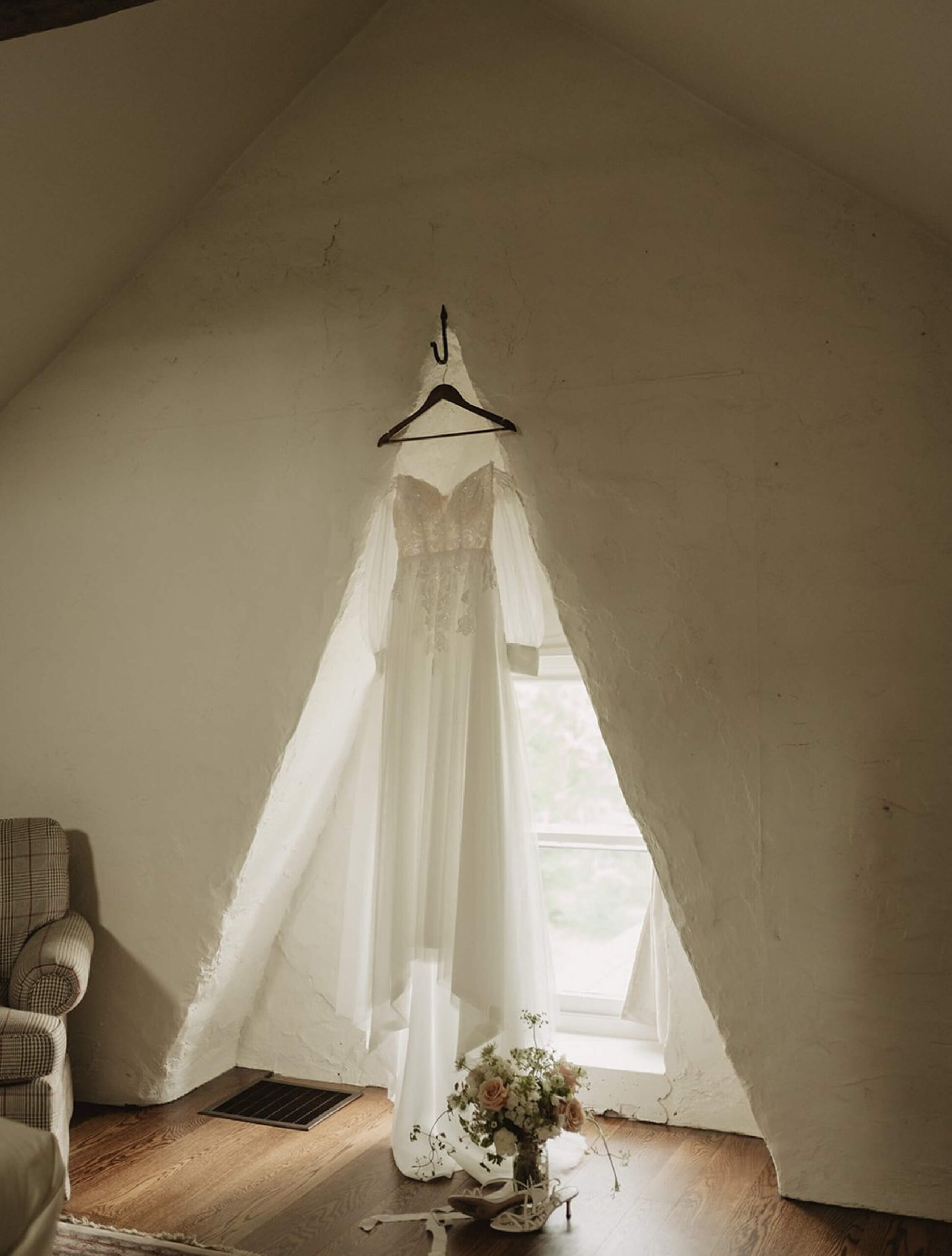 A white wedding dress on a hanger is displayed in front of a small window with a bouquet of flowers on the wooden floor below.
