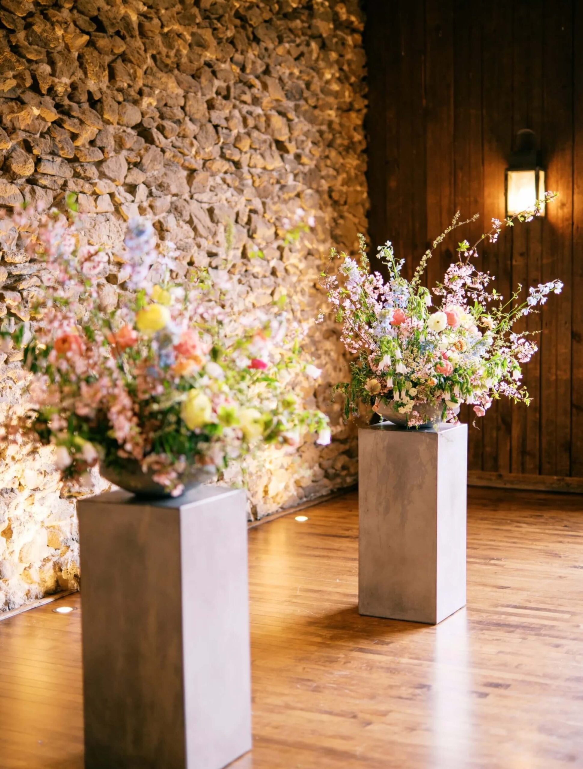 Two large floral arrangements with pastel flowers stand on square concrete pedestals in a room with wooden floors and stone walls.