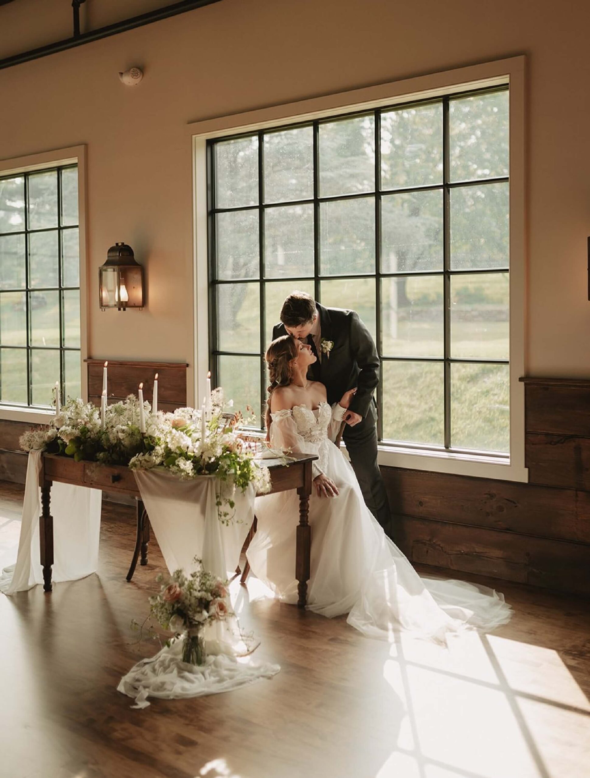 A bride and groom pose together in front of large windows beside a decorated table with flowers and candles in a sunlit room.