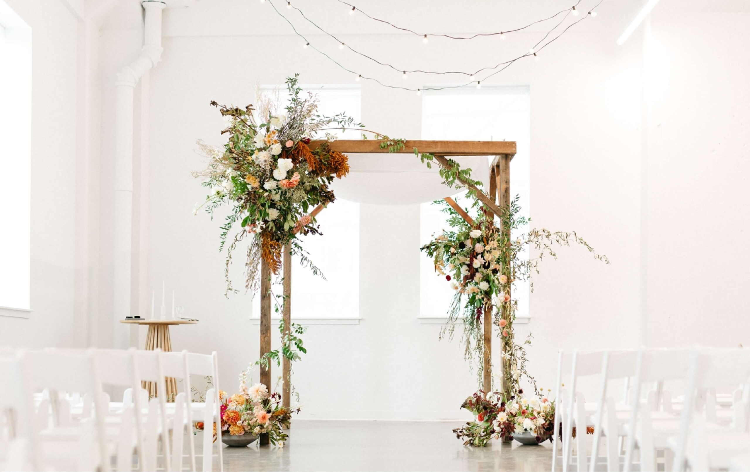 A wooden wedding arch decorated with flowers and greenery stands in a bright, white room with rows of white chairs and string lights overhead.