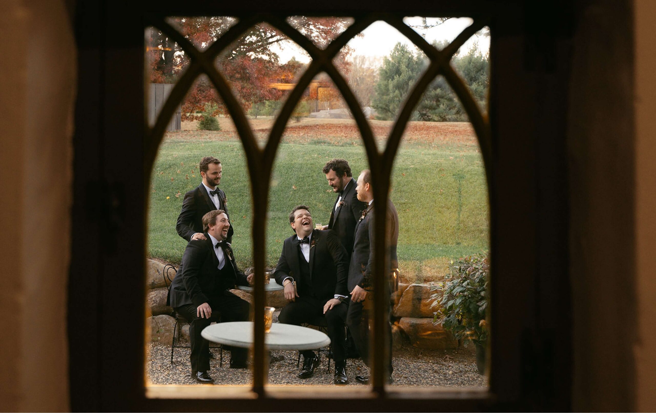 Five men in tuxedos laugh and talk while sitting and standing around a small table outdoors, seen through an arched window.