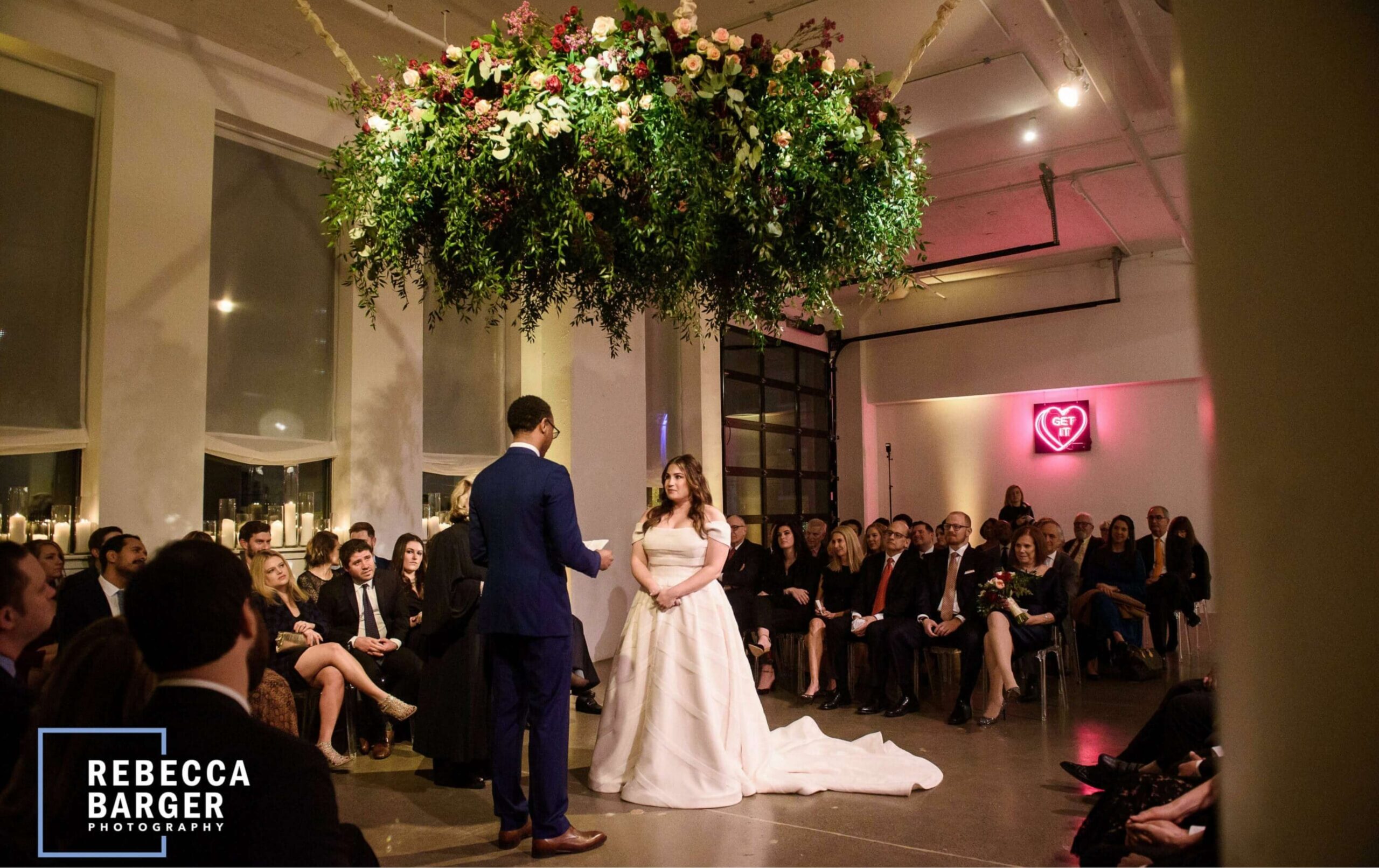 A bride and groom stand together during their wedding ceremony indoors, surrounded by seated guests and greenery hanging overhead.