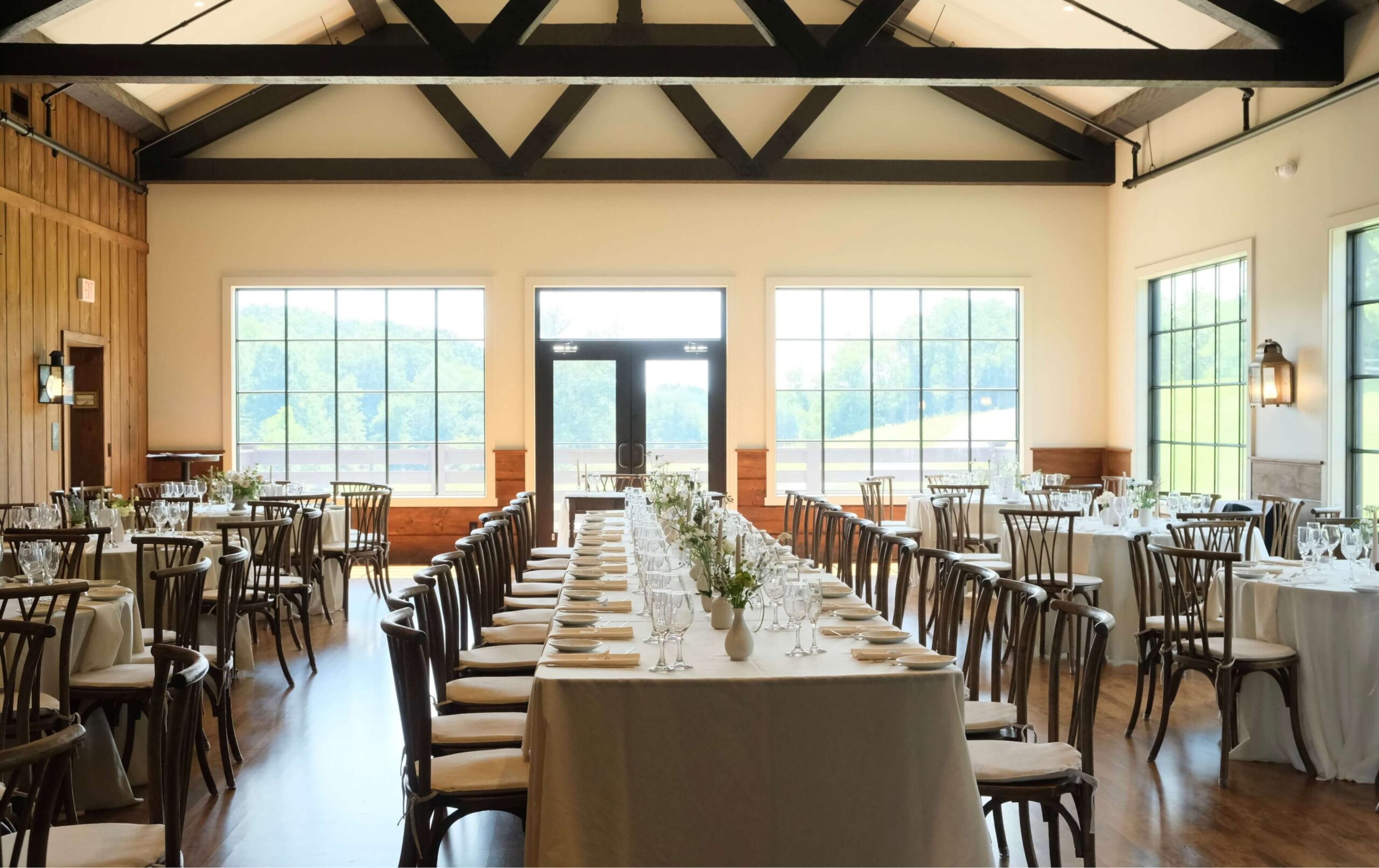 A bright, spacious dining room set for an event with long tables, wooden chairs, white tablecloths, and glassware, illuminated by large windows and natural light.