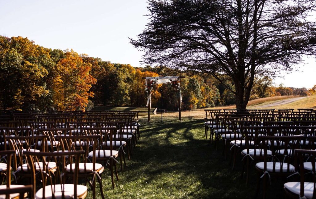 Rows of empty chairs face a simple wedding arch outdoors, surrounded by trees with autumn foliage under clear skies.