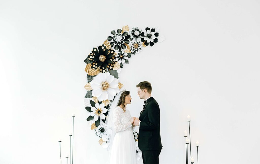 A bride and groom stand facing each other, holding hands, in front of a white wall adorned with large black, white, and gold paper flowers.