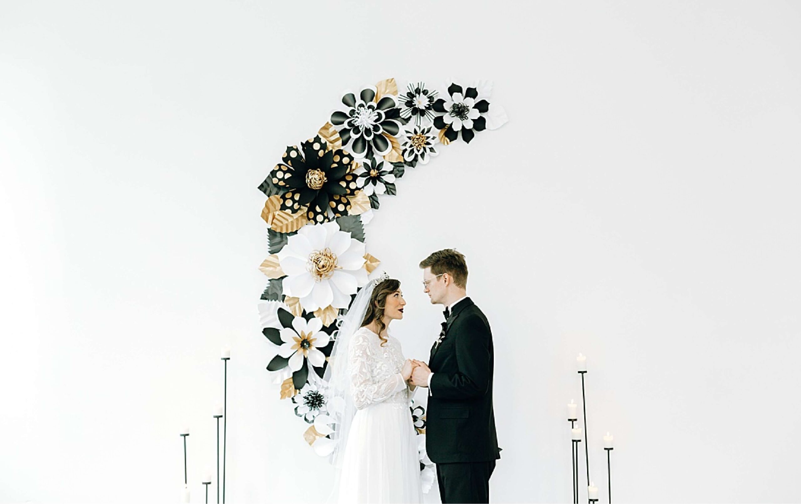 A bride and groom stand facing each other, holding hands, in front of a white wall adorned with large black, white, and gold paper flowers.