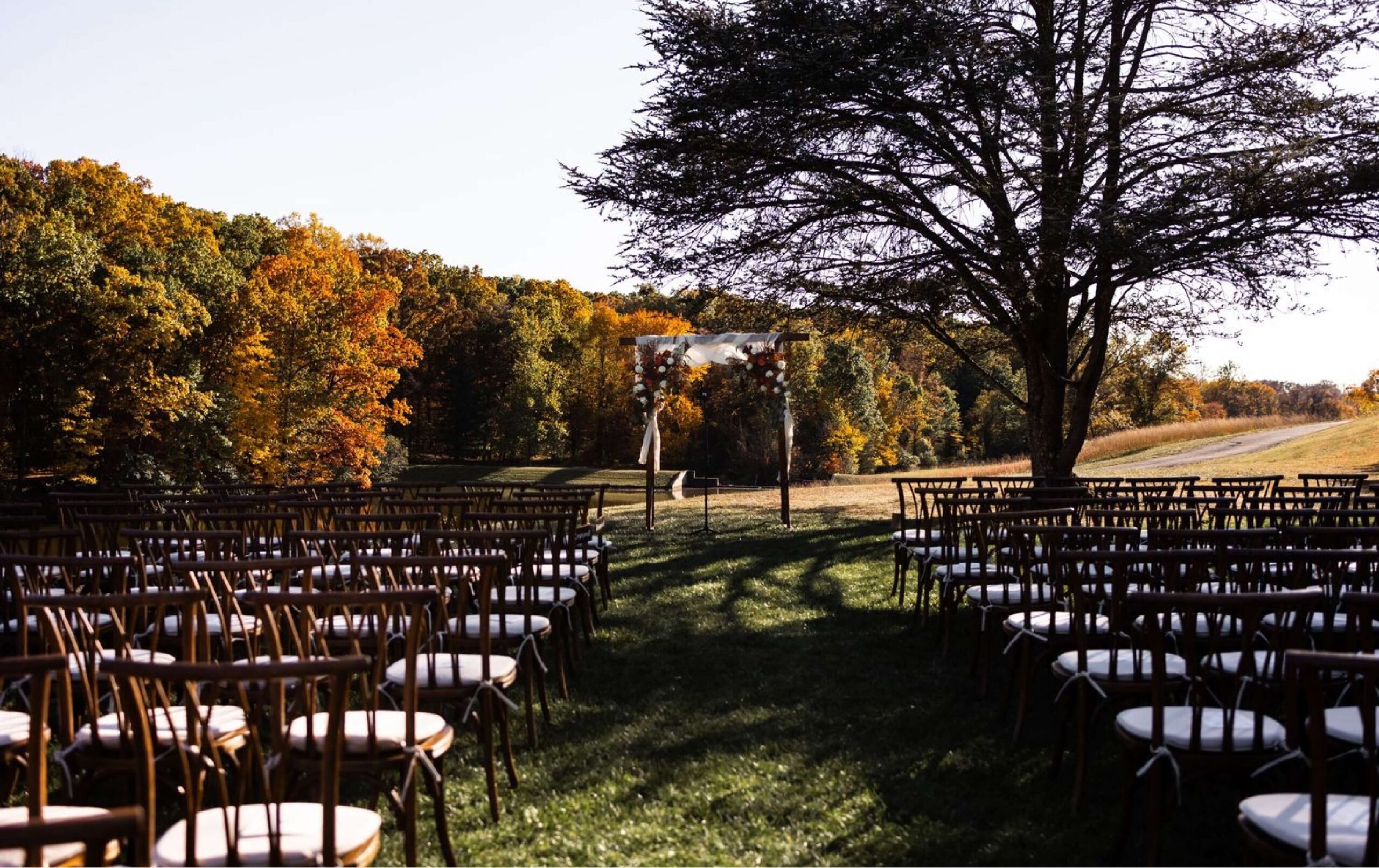 Rows of empty chairs face a simple wedding arch outdoors, surrounded by trees with autumn foliage under clear skies.