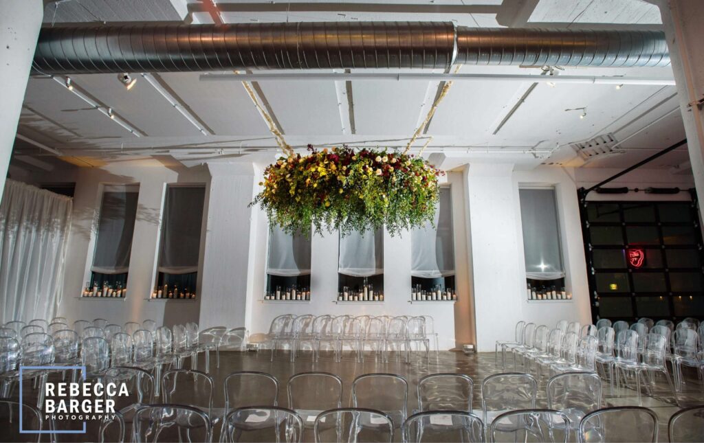 Modern indoor event space with transparent chairs, a large suspended floral arrangement, and candles lining windowsills; industrial ceiling details visible.