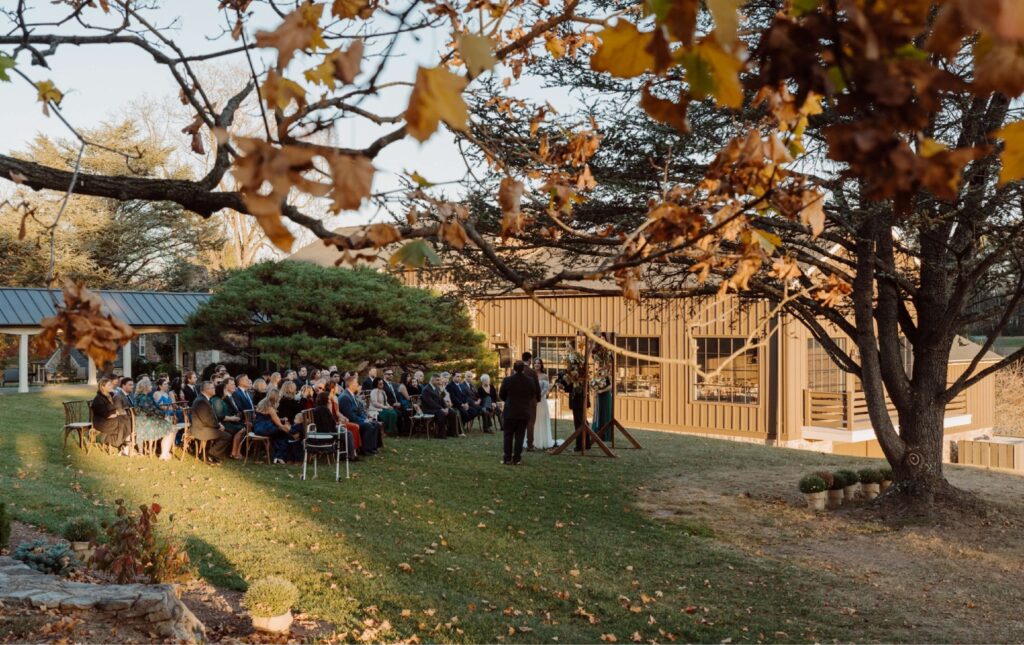 A group of people sit outdoors on a lawn, attending a wedding ceremony near a building, with autumn trees and fallen leaves in the foreground.