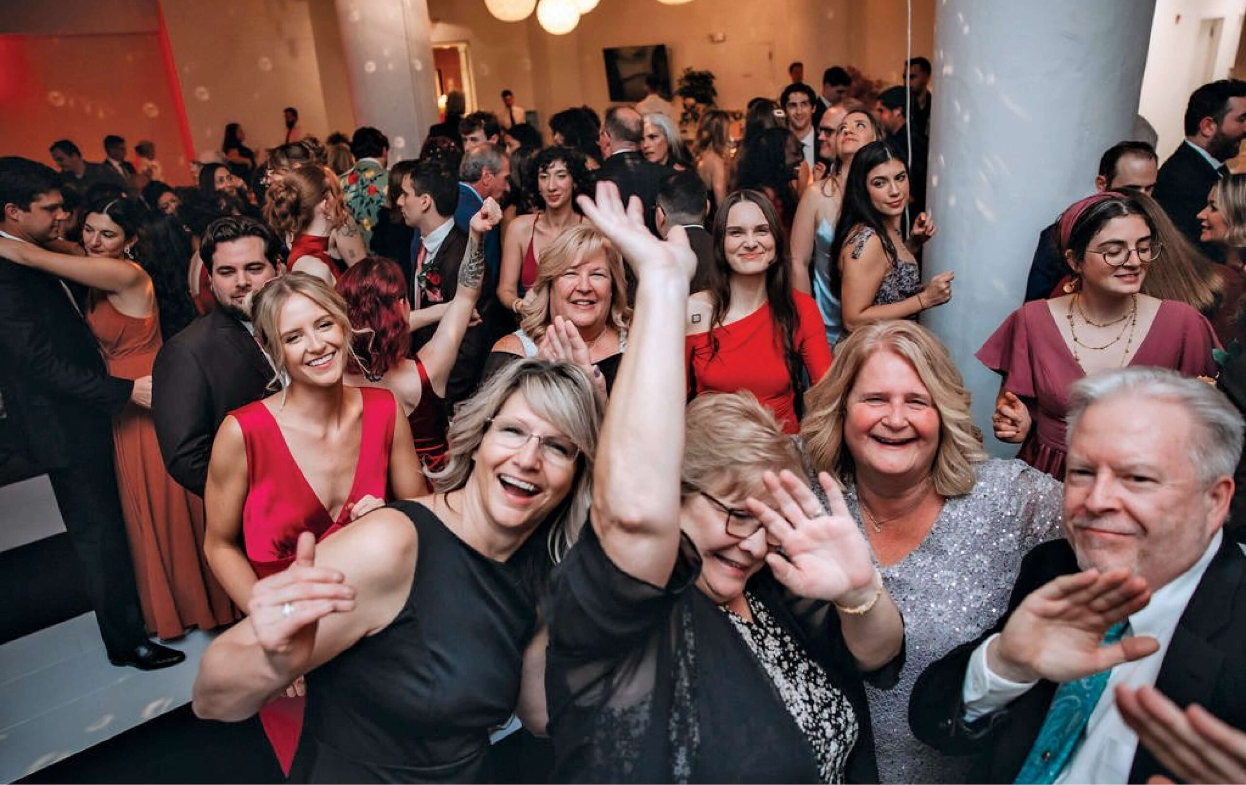 A large group of people dressed formally are smiling, dancing, and waving at a lively indoor party or celebration.