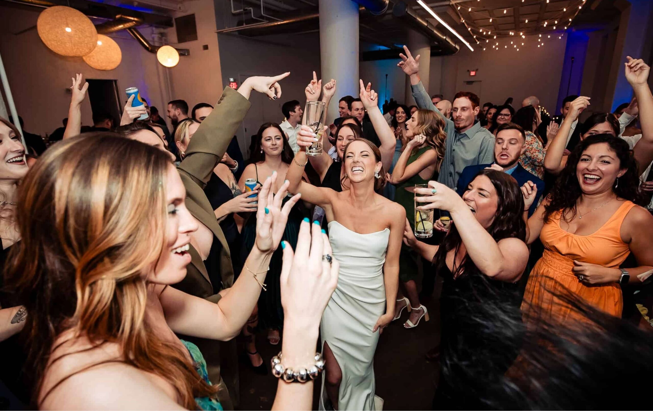 A group of people, including a woman in a white dress, dance and celebrate together at an indoor party or wedding reception.