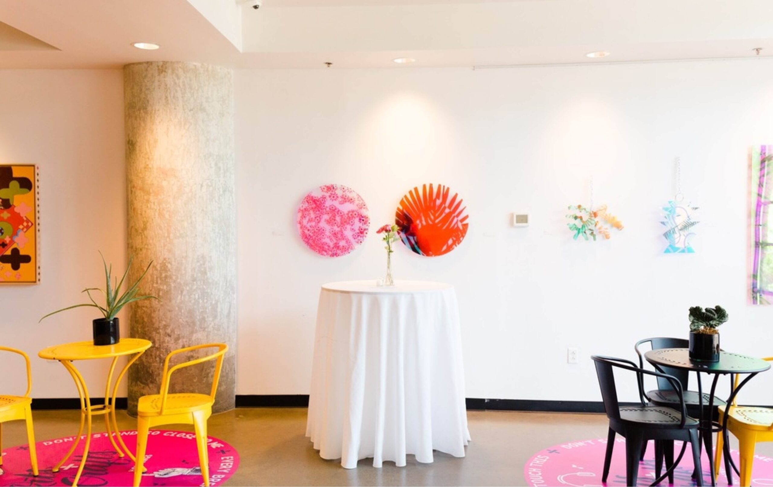 Gallery-style room with colorful abstract art on the white wall, a round table with a white tablecloth and vase, and yellow and black chairs with small potted plants.
