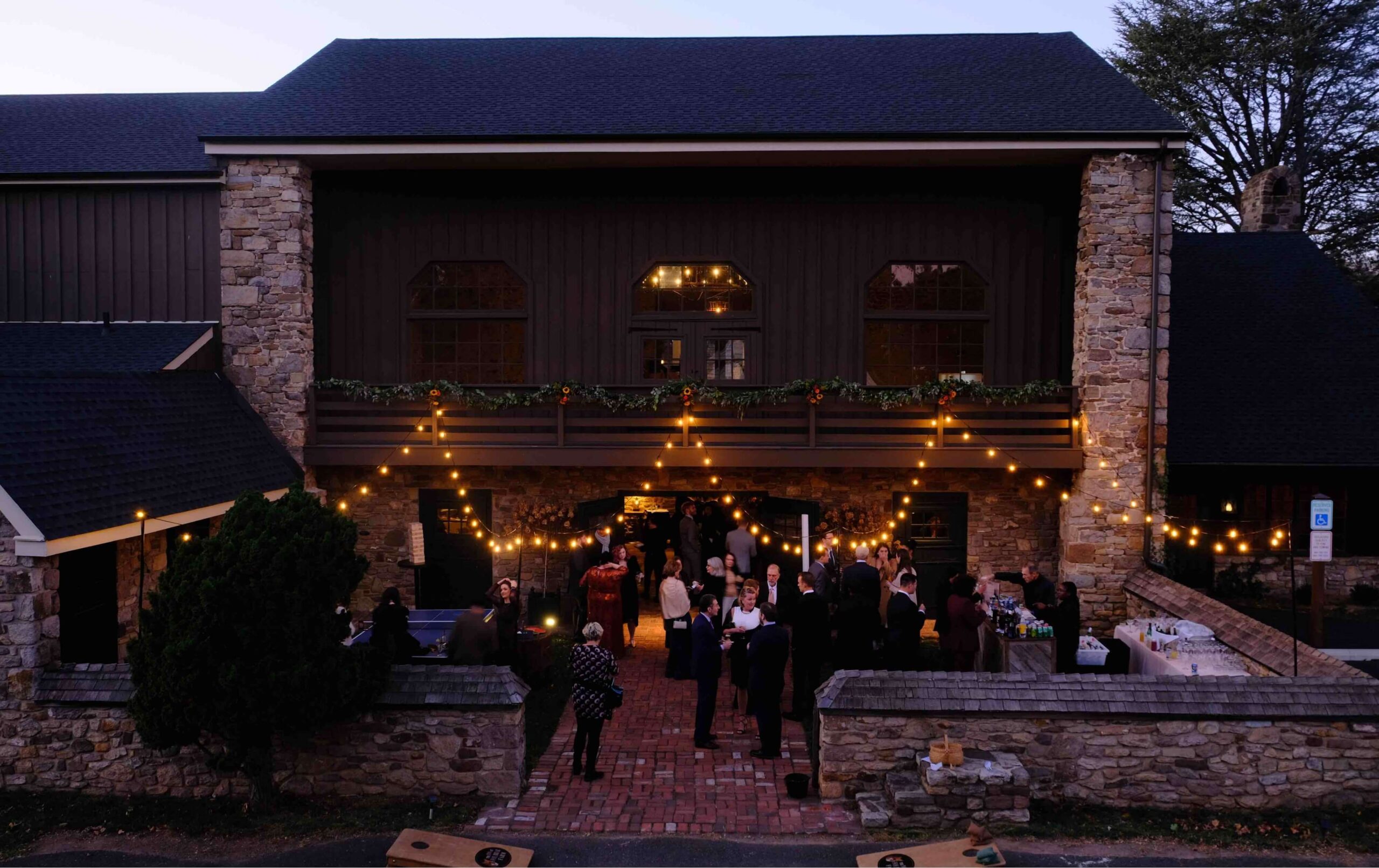 A group of people gathered outside a stone building at dusk, under string lights, with some playing lawn games and others socializing near the entrance.