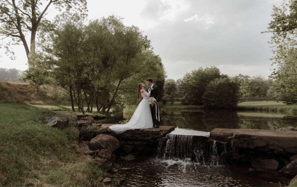 A bride and groom stand together on a stone bridge over a small waterfall, surrounded by trees and greenery near a calm pond.