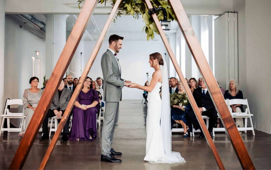 A bride and groom stand holding hands under a wooden triangular arch during a wedding ceremony, with seated guests watching in a modern indoor venue.