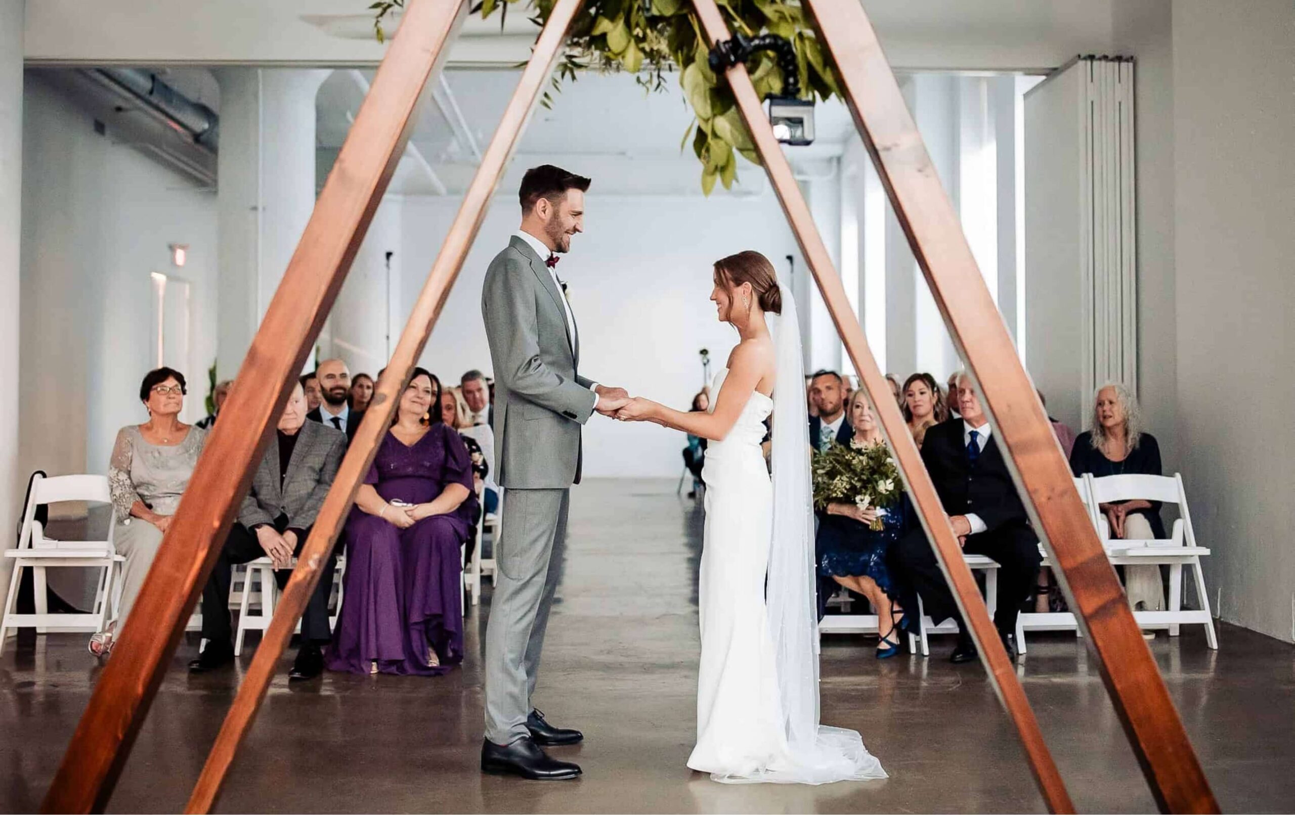A bride and groom stand holding hands under a wooden triangular arch during a wedding ceremony, with seated guests watching in a modern indoor venue.