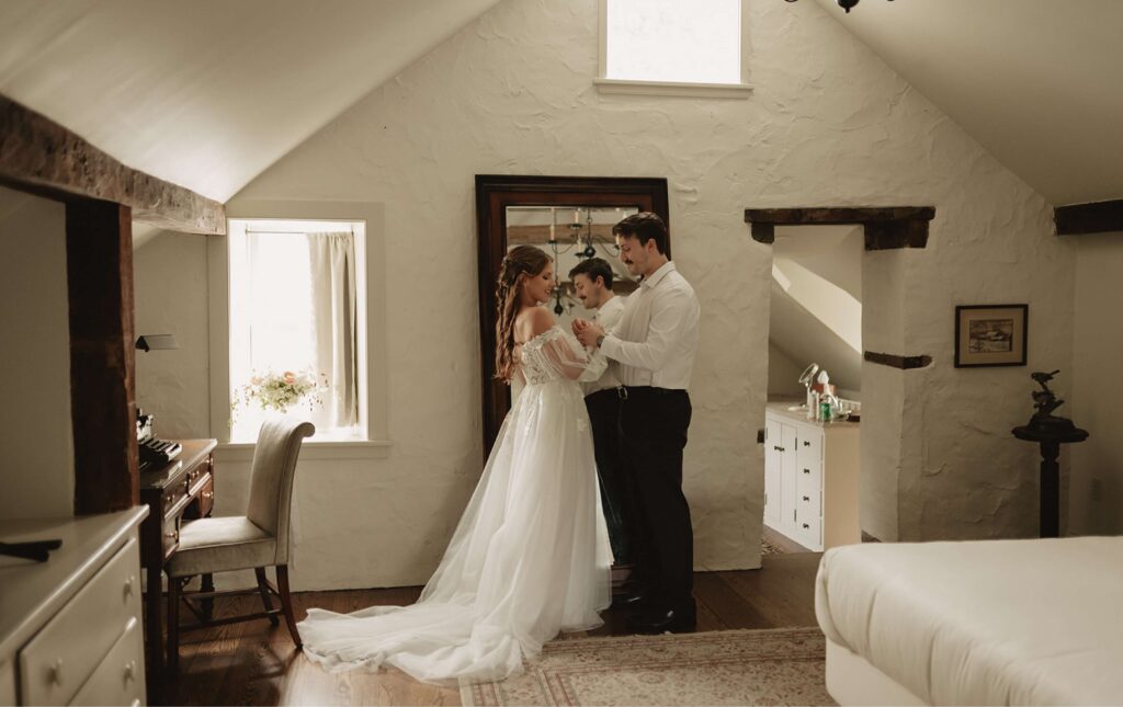 A bride and groom stand in a softly lit room, holding a young child between them, with vintage decor and natural light streaming through the windows.