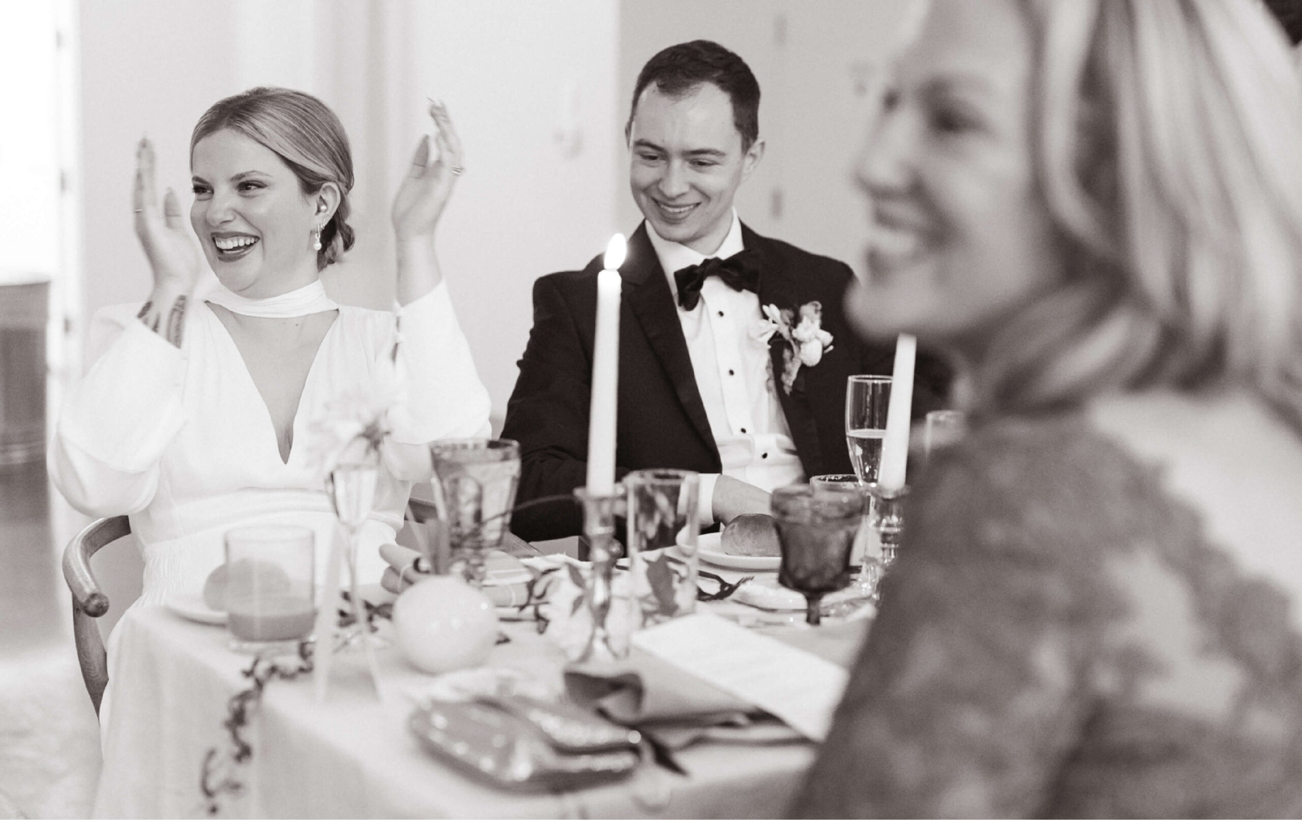 A bride and groom sit at a decorated table with a guest, all dressed formally; the bride is smiling with hands raised in a celebratory gesture.