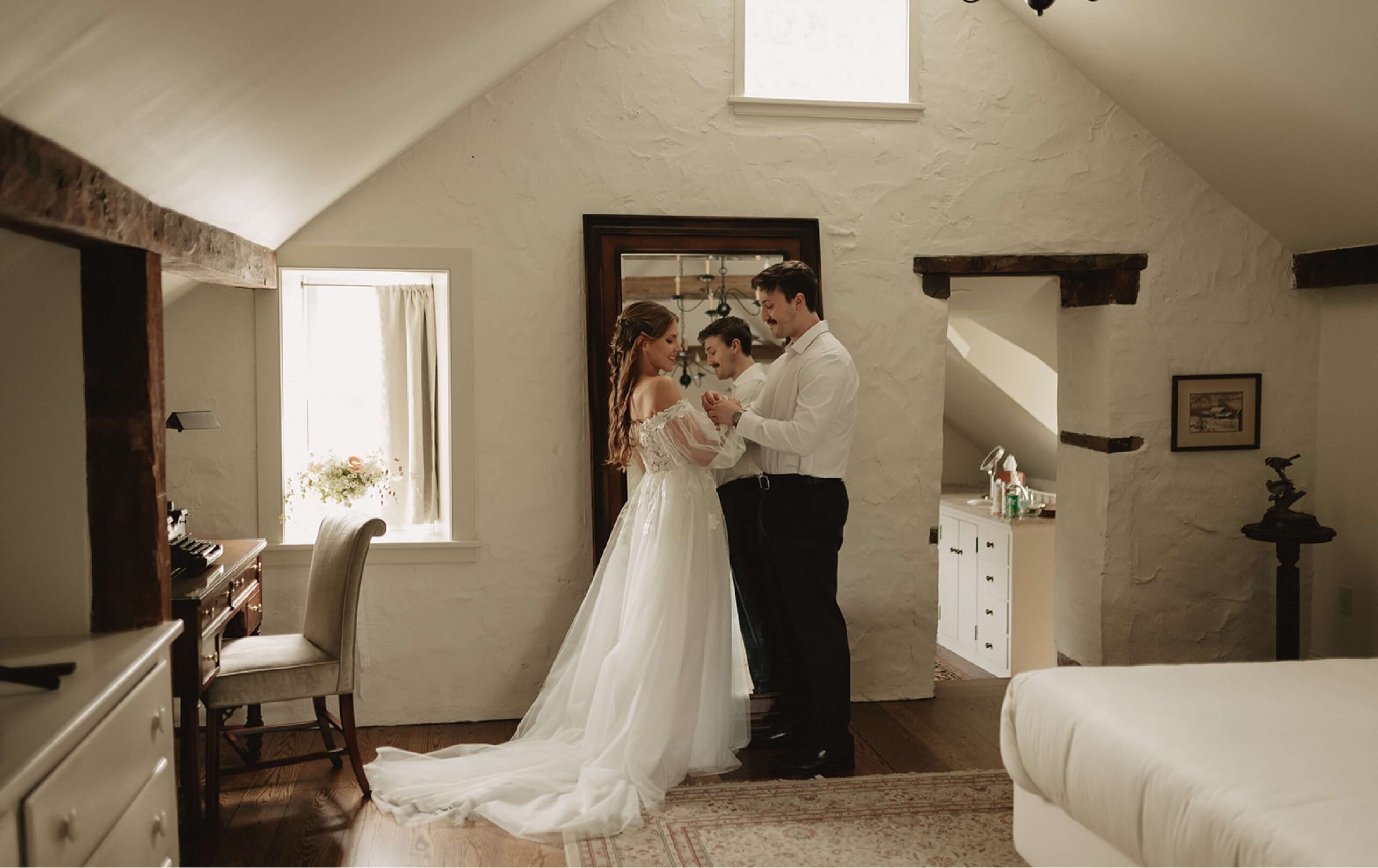 A bride and groom stand in a softly lit room, holding a young child between them, with vintage decor and natural light streaming through the windows.