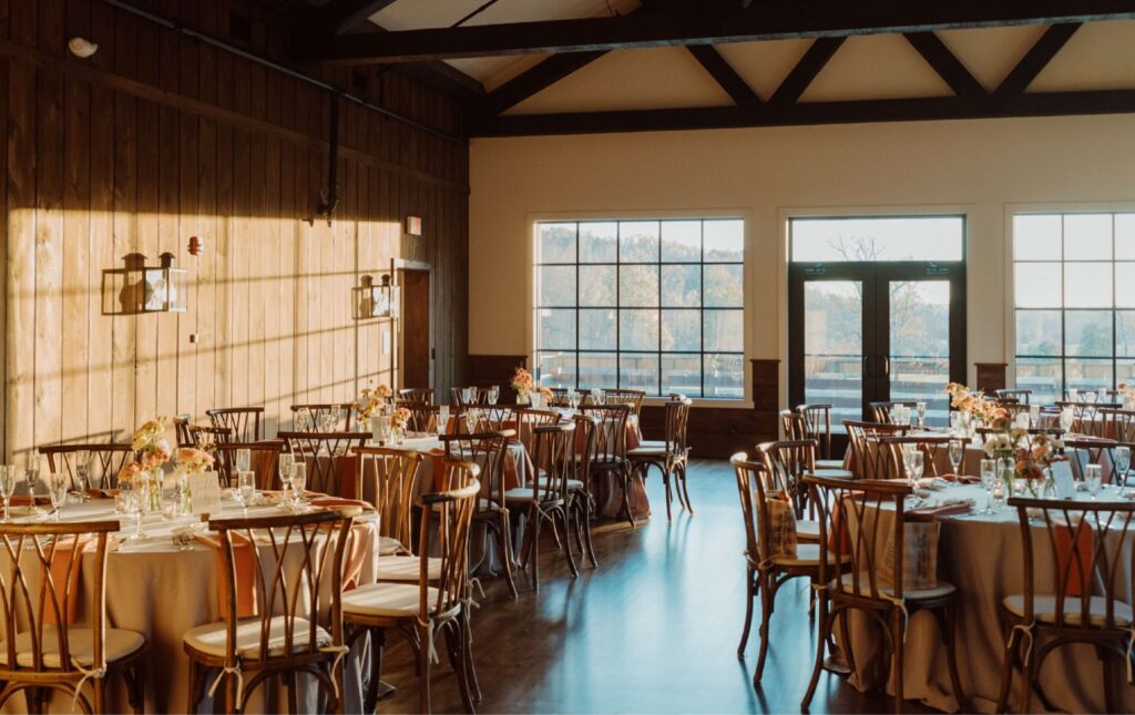 A sunlit banquet hall with round tables set for an event, wooden chairs, floral centerpieces, and large windows letting in natural light.