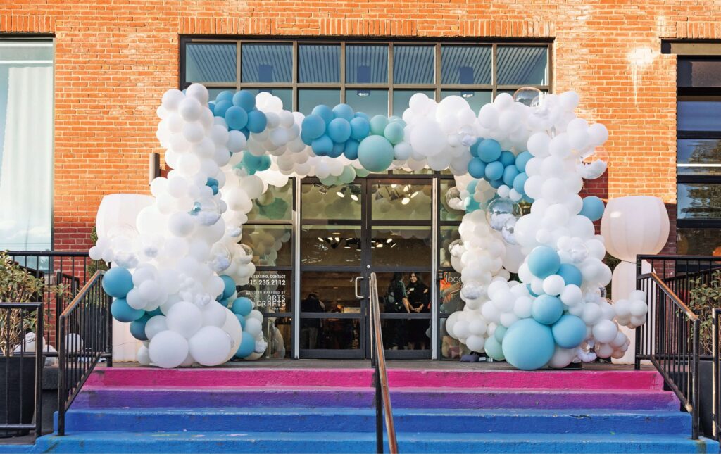 A brick building entrance decorated with a large balloon arch in white and blue, above glass double doors and pink to blue gradient-painted steps.