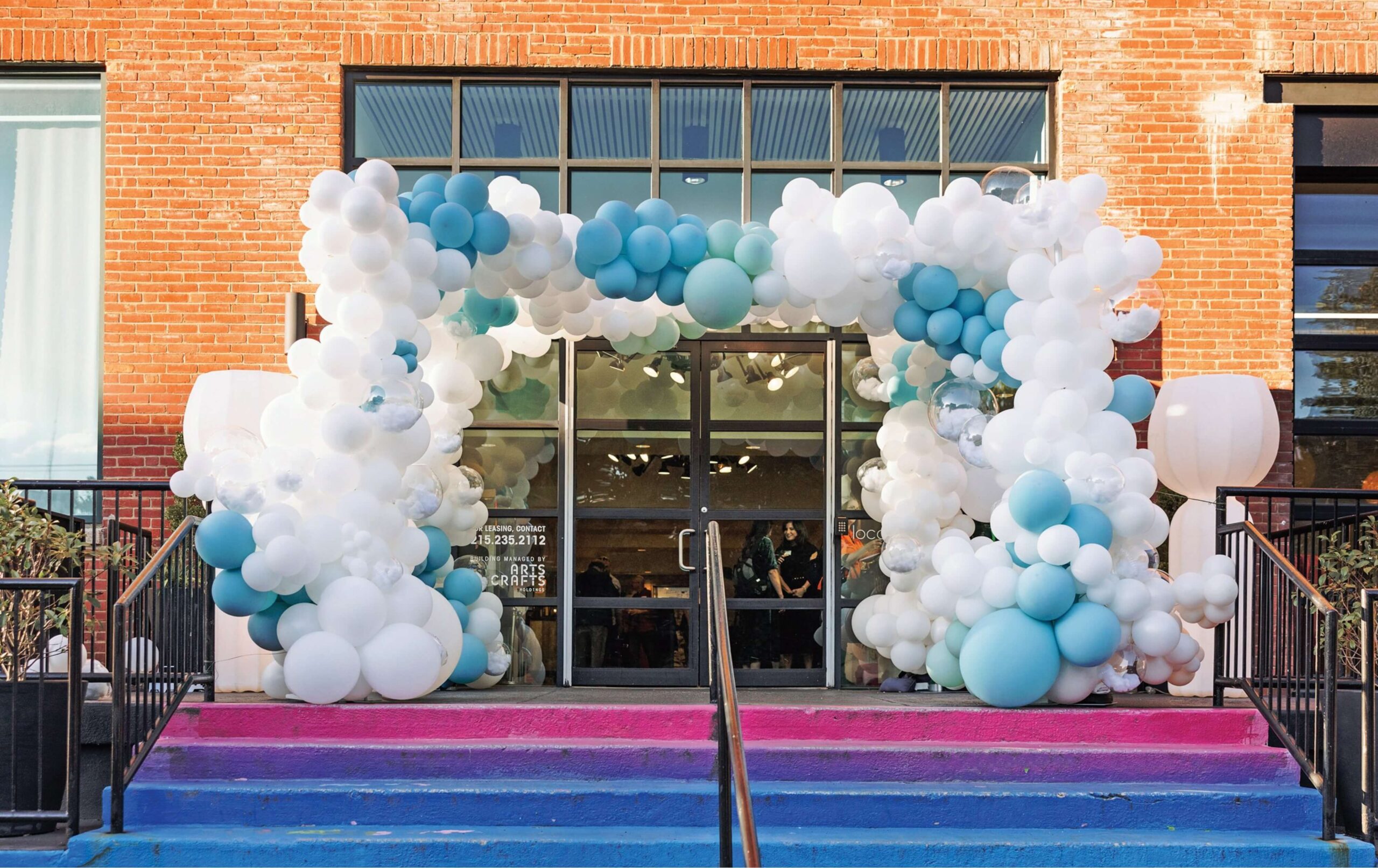 A brick building entrance decorated with a large balloon arch in white and blue, above glass double doors and pink to blue gradient-painted steps.