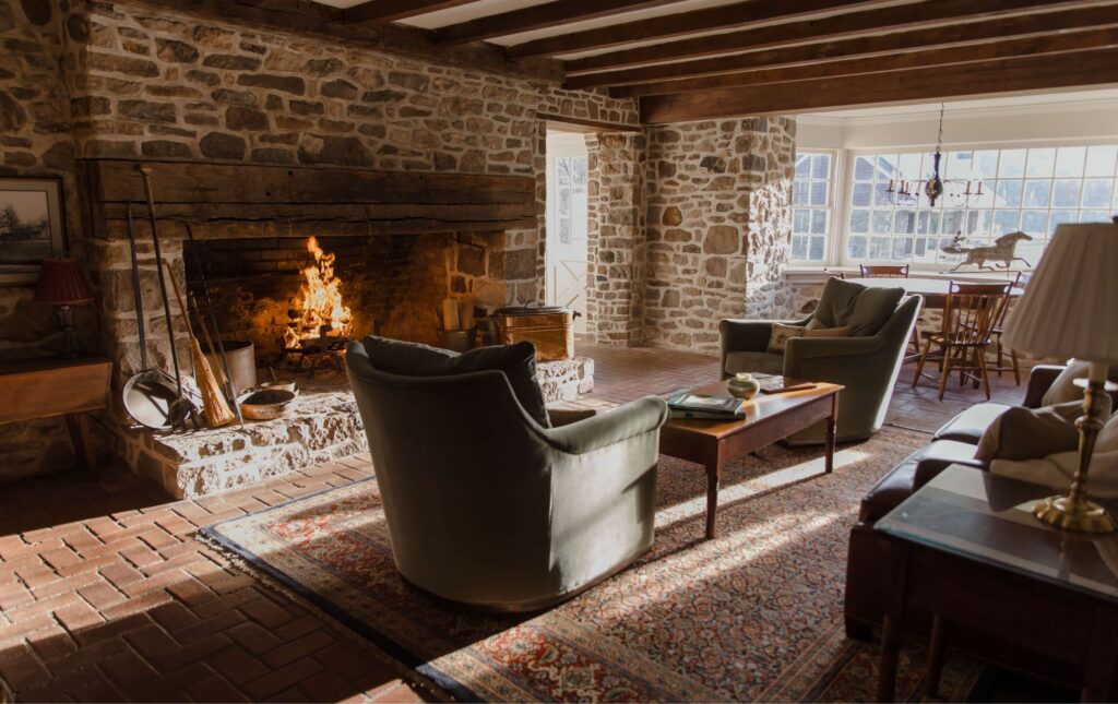 A stone-walled living room features a lit fireplace, two armchairs, a wooden coffee table, and large windows letting in sunlight.