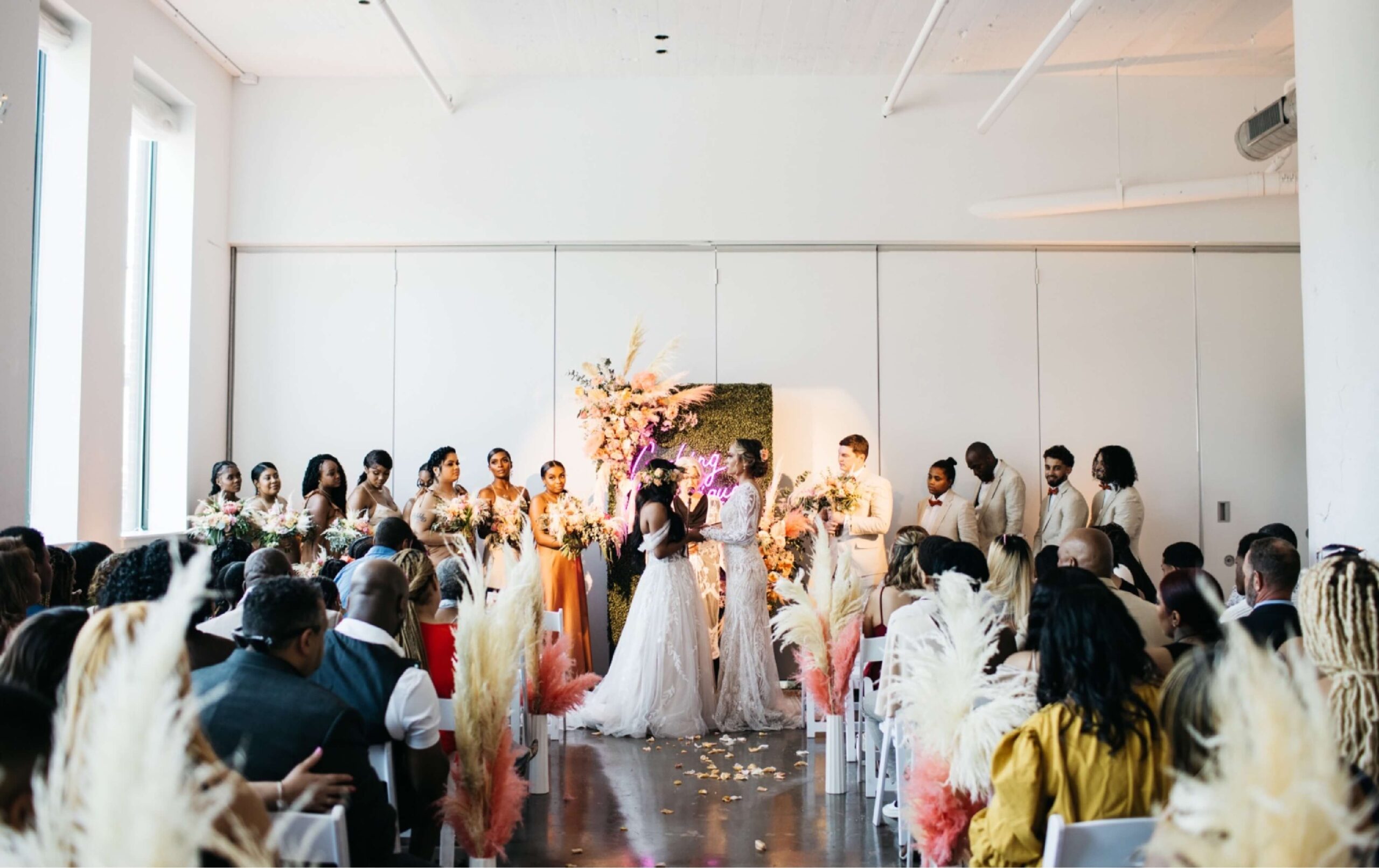 A bride and groom stand at the altar with their wedding party on either side, facing seated guests in a bright, decorated indoor venue.