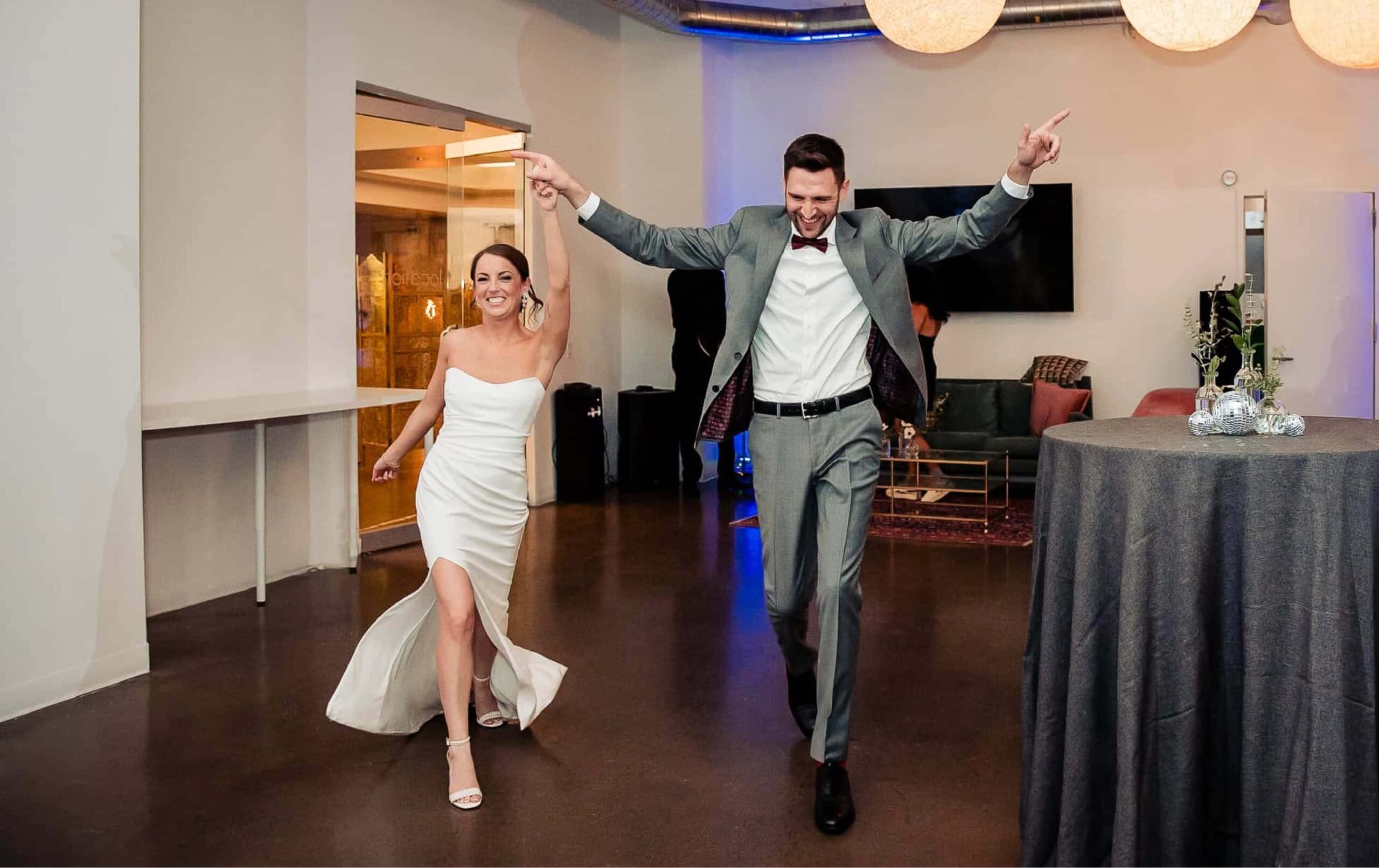 A woman in a white dress and a man in a gray suit enter a room smiling, holding hands, and raising their arms in celebration.